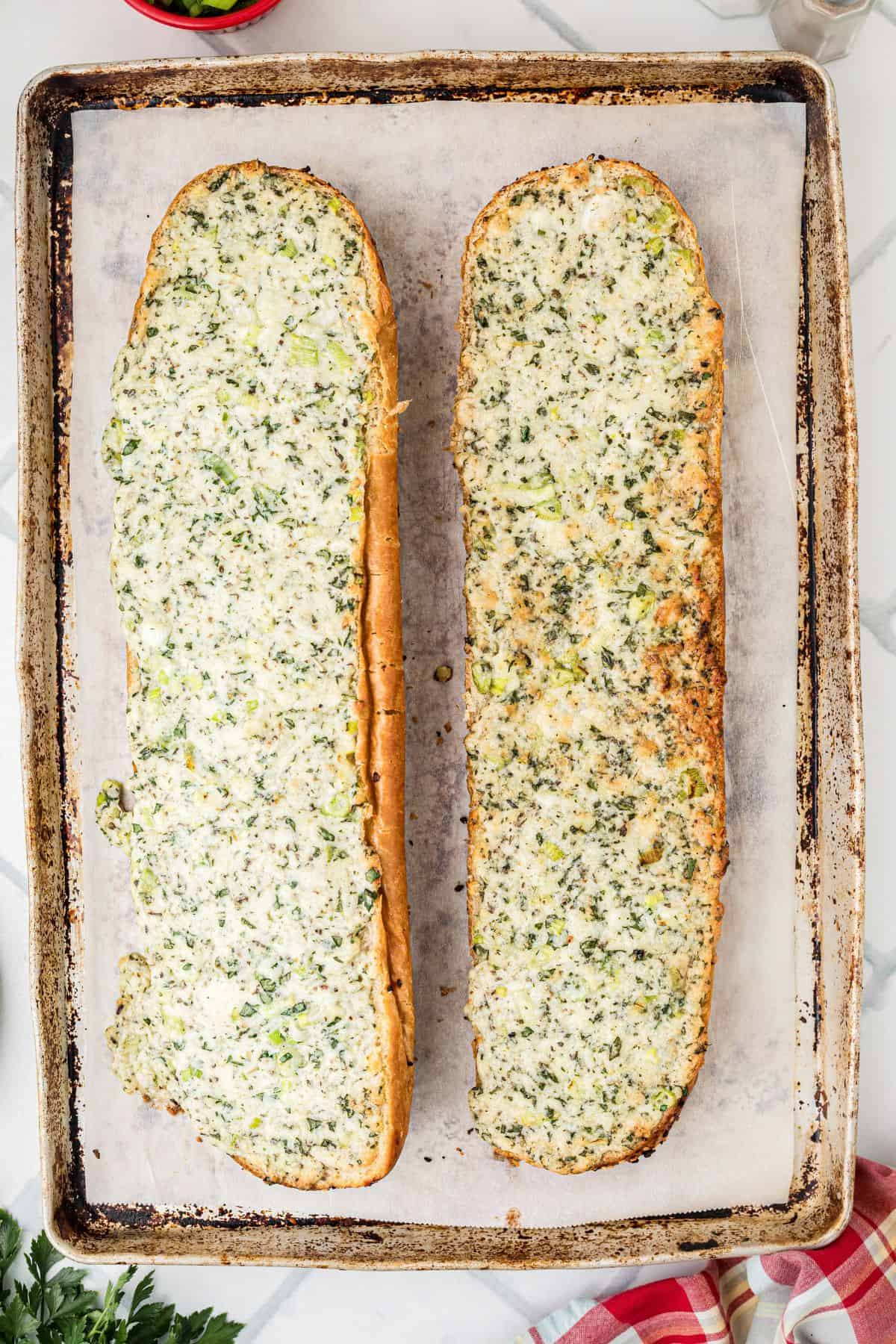 A baking sheet lined with parchment paper holds two halves of a loaf of garlic bread topped with melted cheese and herbs. The bread is golden brown around the edges.