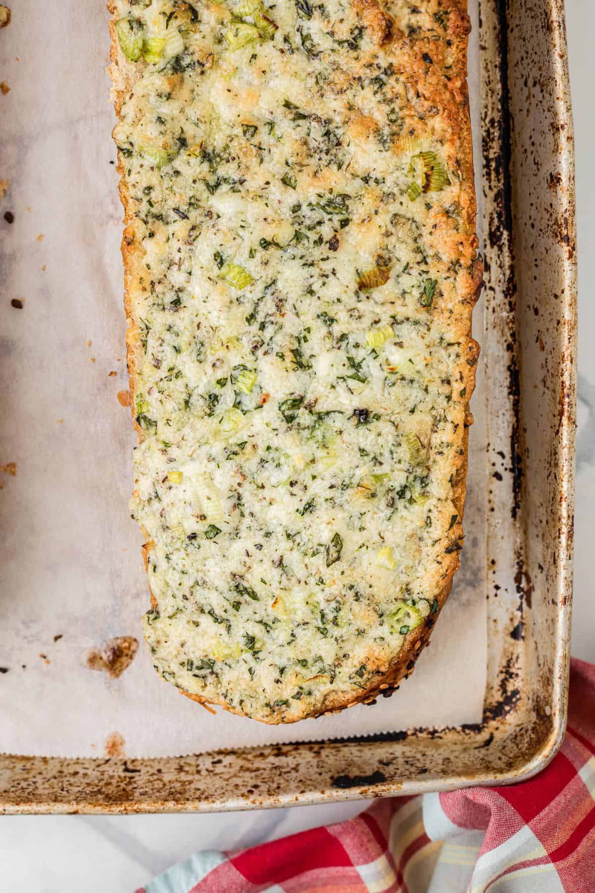 A close-up of a piece of garlic bread with herbs on a parchment-lined baking sheet. The bread is golden brown around the edges, and a red-and-white checkered cloth is partially visible in the lower right corner.