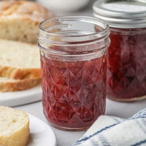 A small jar of strawberry jam with toast in the background.