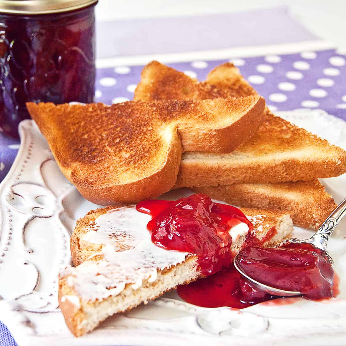 Strawberry jam on buttered toast with jars of jam in the background.
