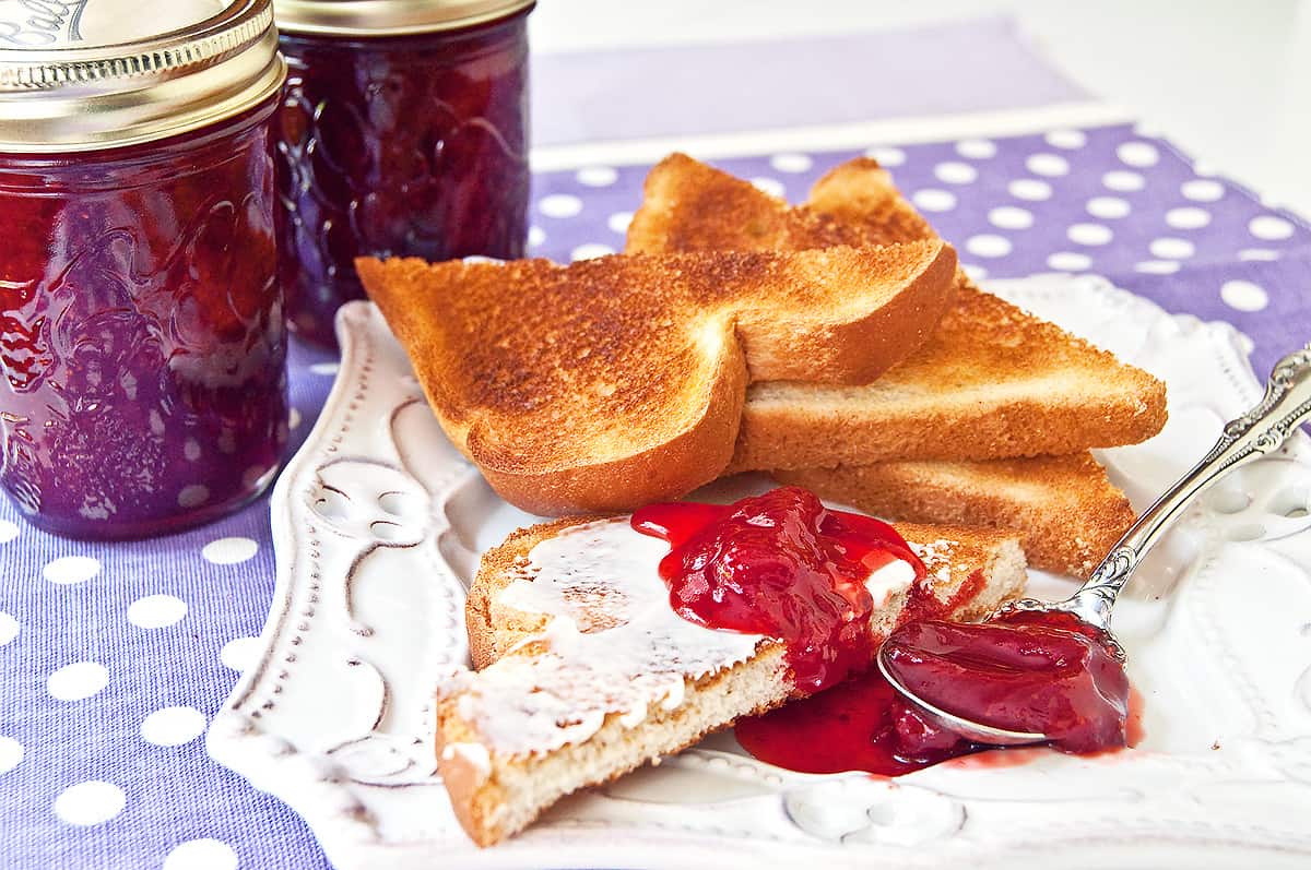 Strawberry jam on buttered toast with jars of jam in the background.