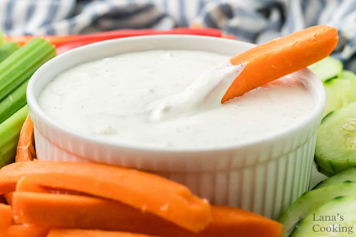 Carrot stick dipped in blue cheese dip in a white bowl, surrounded by fresh vegetables.