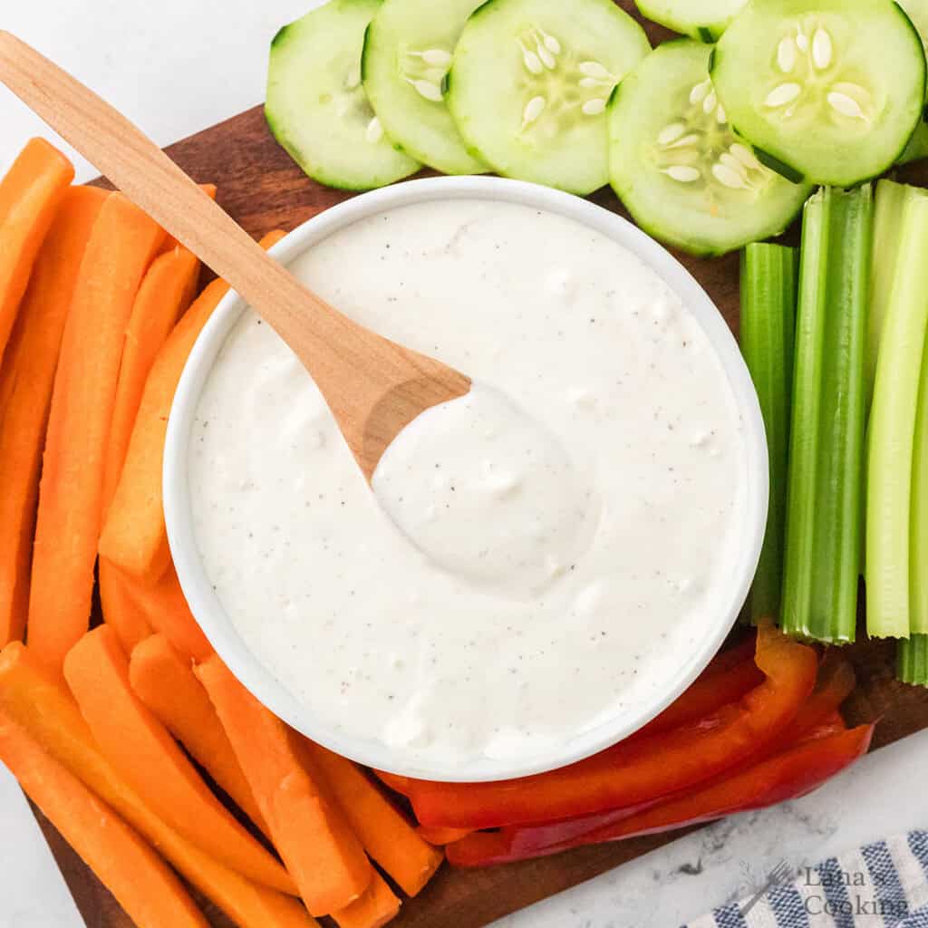 Bowl of blue cheese dip with a wooden spoon, surrounded by sliced carrots, celery, cucumber, and bell peppers.