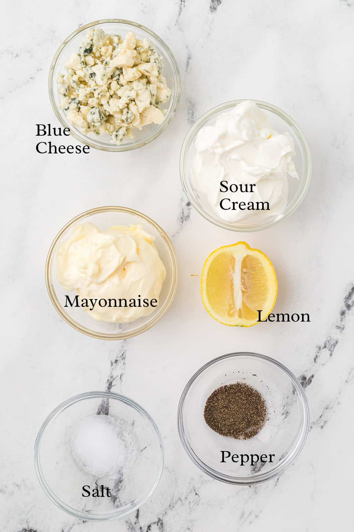 Five bowls labeled blue cheese, sour cream, mayonnaise, salt, pepper, plus half a lemon, on white marble.