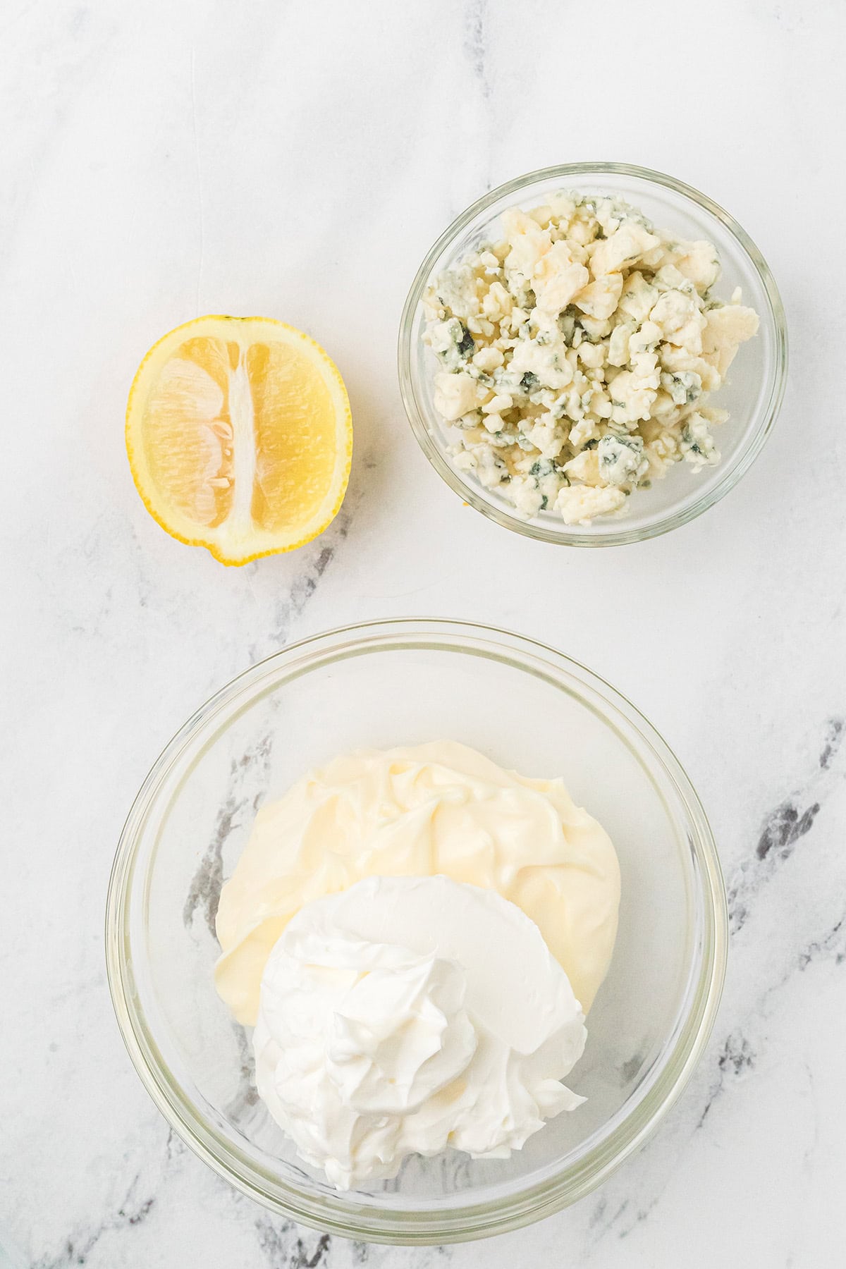 A bowl of mayo and sour cream, a bowl of crumbled blue cheese, and half a lemon on a white surface.