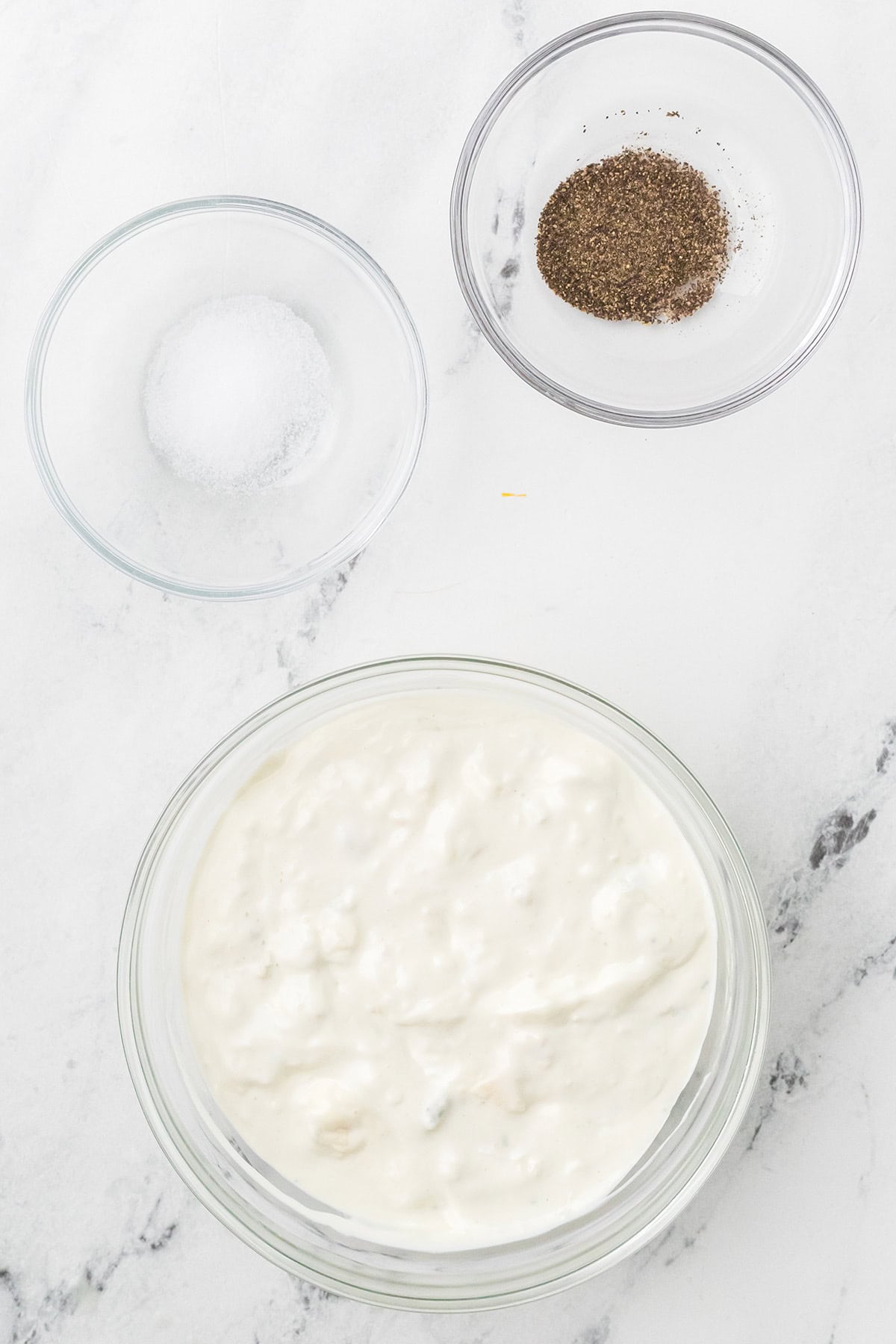 Three glass bowls: one with white dip, one with salt, and one with ground black pepper on a marble surface.