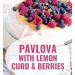 Pavlova topped with lemon curd, raspberries, blueberries, and lemon zest on a glass cake stand.