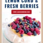 A pavlova topped with lemon curd, raspberries, blueberries, and lemon zest on a glass cake stand.