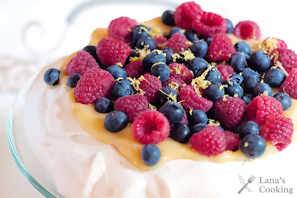 A pavlova topped with fresh raspberries, blueberries, lemon zest, and a layer of golden curd.