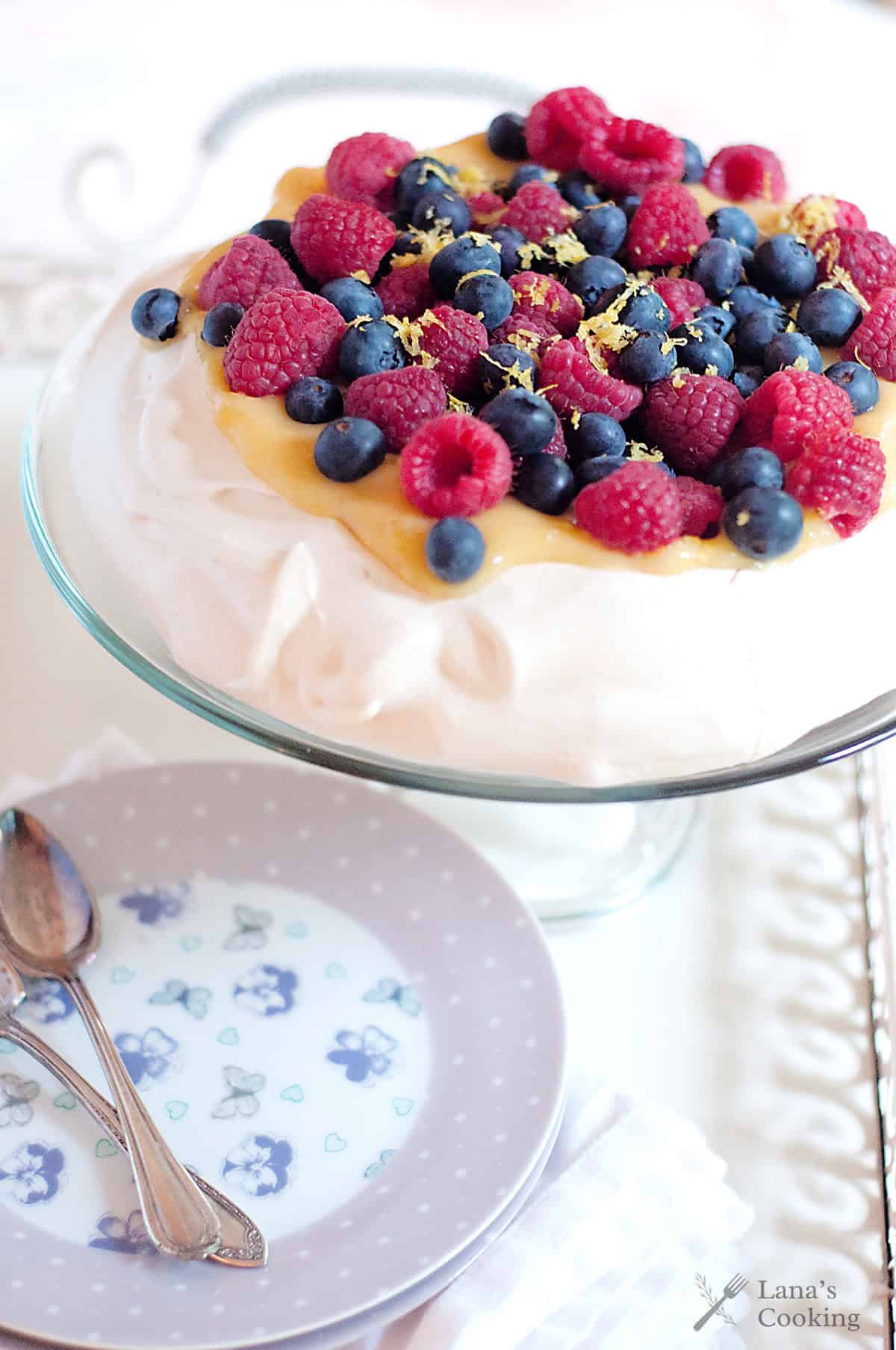 A pavlova topped with lemon curd, raspberries, and blueberries on a glass stand next to a floral plate and spoons.