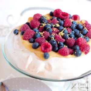 A pavlova topped with lemon curd, fresh raspberries, blueberries, and lemon zest on a glass stand.