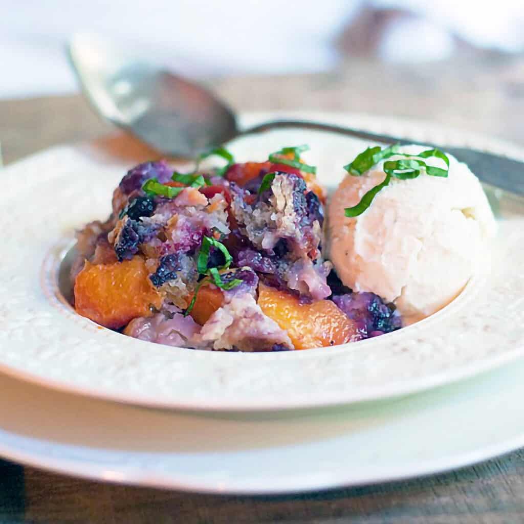 A serving of peach blueberry basil cobbler in a bowl with ice cream.