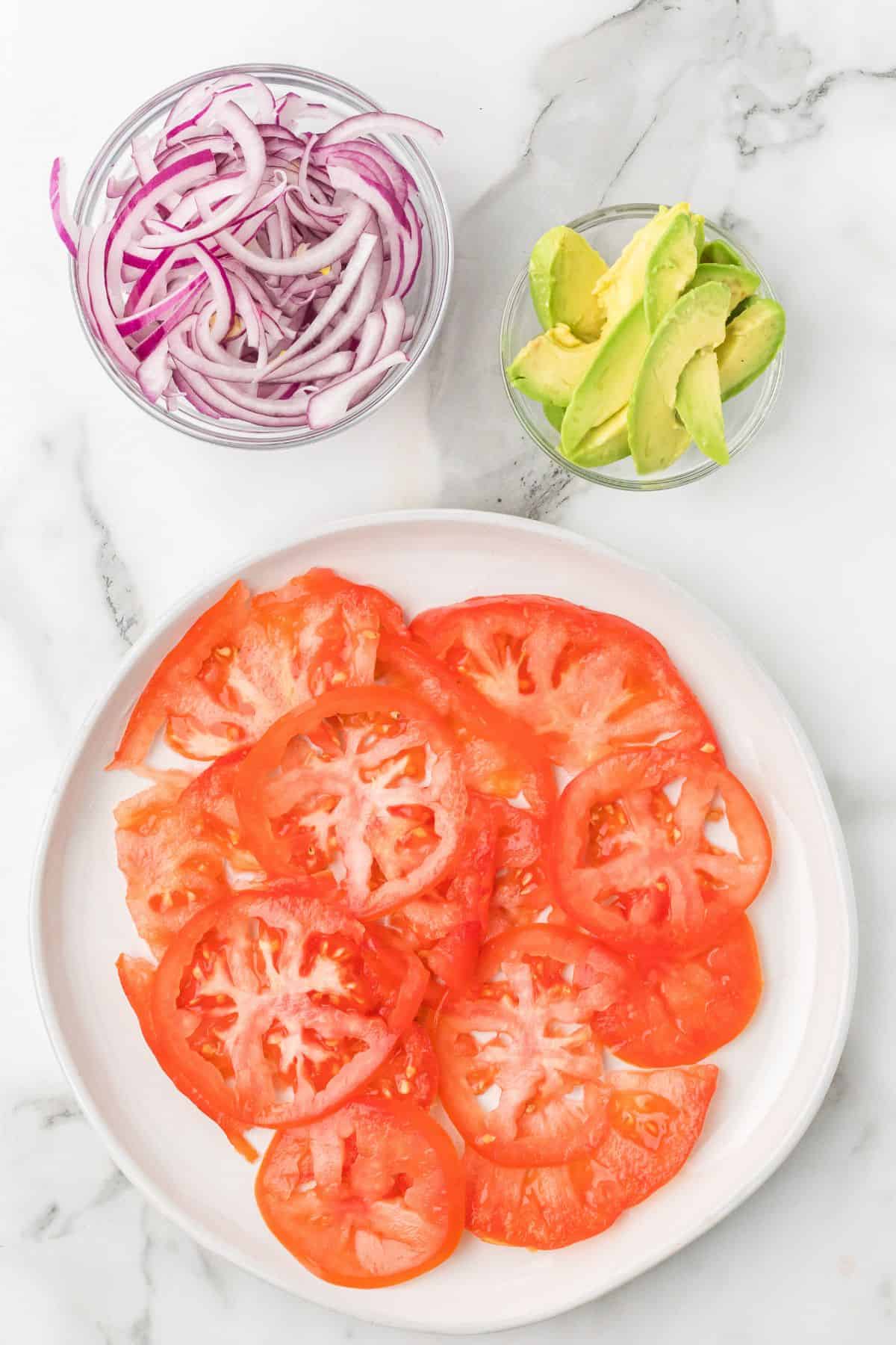 Sliced tomatoes on a plate, with bowls of sliced red onions and avocado on a marble surface.
