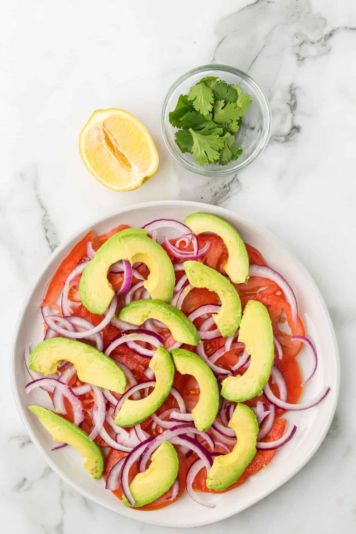 A plate of sliced avocado, tomato, and red onion with a lemon wedge and cilantro on the side.