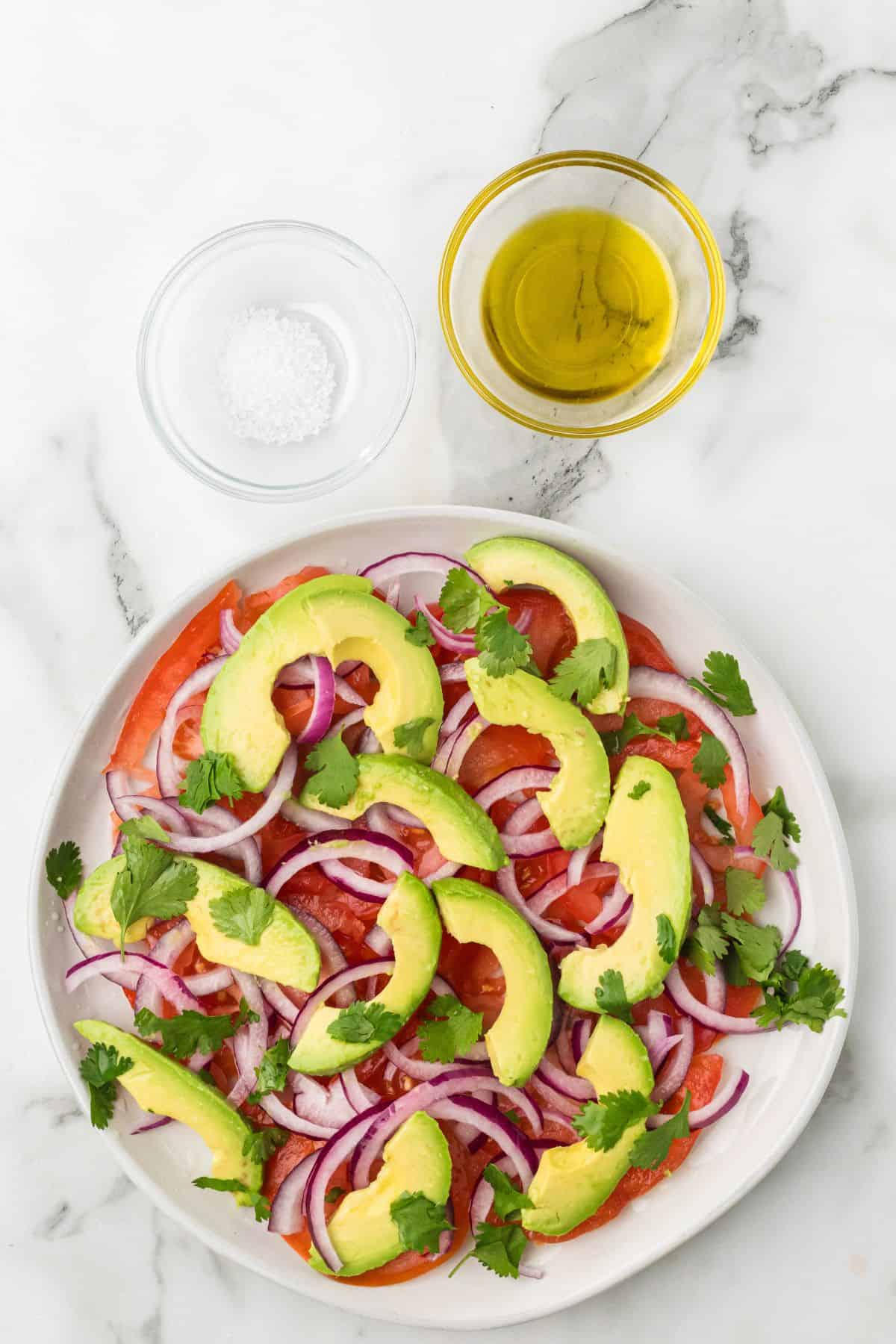 Plate of tomato and avocado salad with red onion and cilantro, alongside bowls of salt and olive oil.