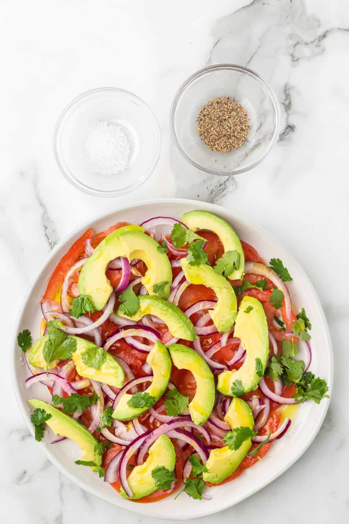 A plate of vibrant tomato and avocado salad with red onion and cilantro sits next to bowls of salt and pepper on marble.