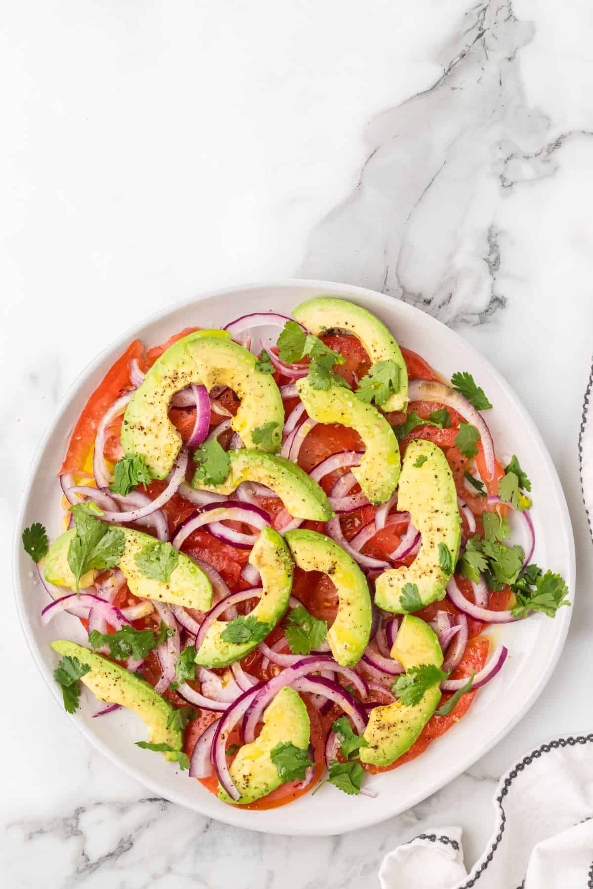 A plate of tomato and avocado salad with sliced avocado, tomato, red onion, and cilantro on a white marble surface.