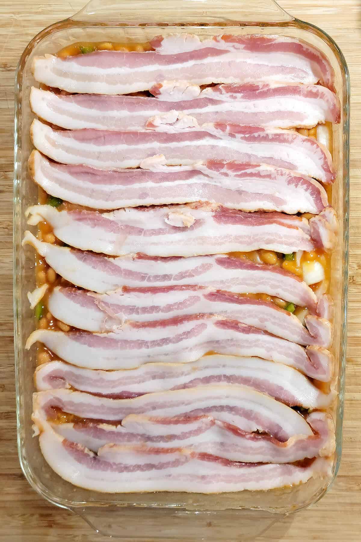 Baked beans in a glass dish covered with bacon strips.
