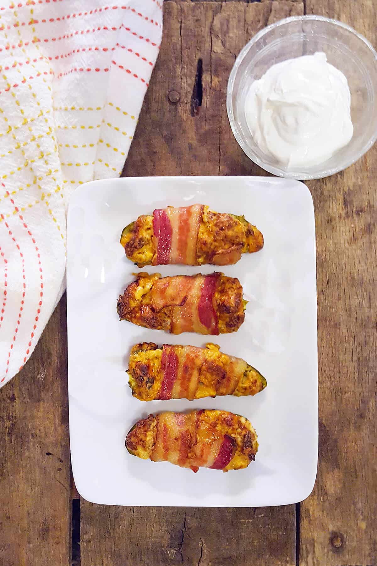 Sausage stuffed bacon wrapped jalapenos on a white serving plate with a kitchen towel and small bowl of sour cream.