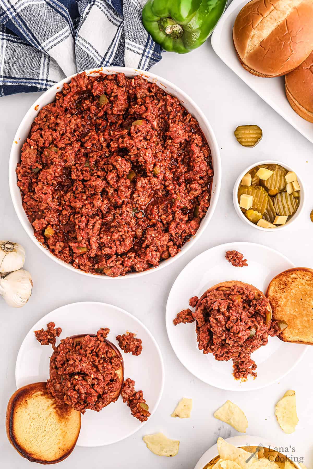 Overhead view of sloppy joe meat in a bowl, buns, pickles, chips, and ingredients on a white surface.