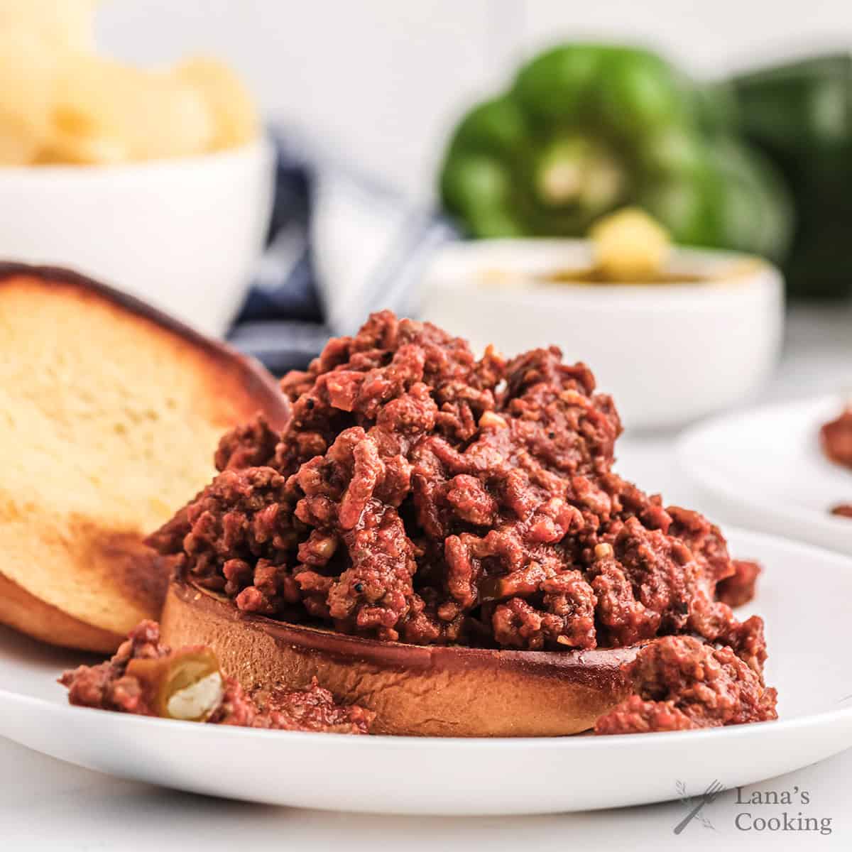 An open-faced sloppy joe sandwich with ground beef filling on a toasted bun, served on a white plate with blurred vegetables and chips in the background.