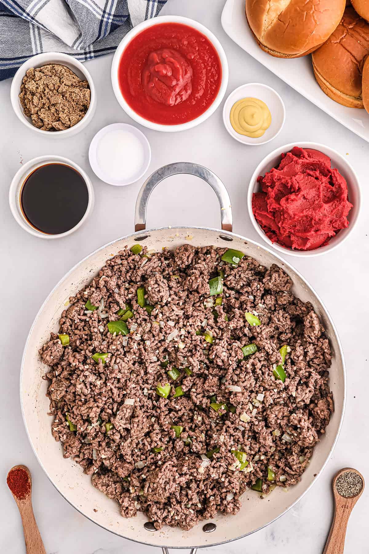 Top-down view of a skillet with cooked ground beef and chopped green onions, surrounded by bowls of sauces, condiments, seasonings, and hamburger buns on a white surface.