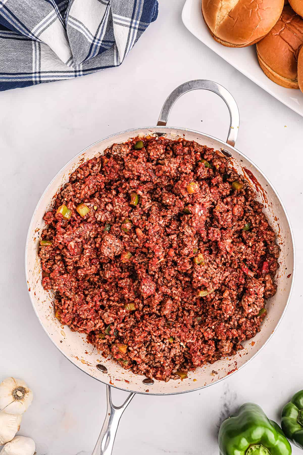 A skillet filled with cooked sloppy joe mixture sits on a white countertop next to hamburger buns, a blue plaid napkin, garlic, and green bell pepper.