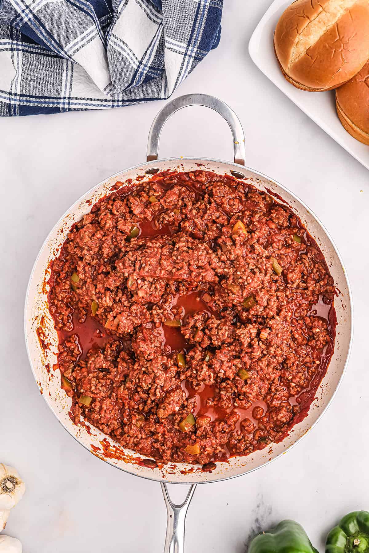 A skillet filled with cooked sloppy joe meat mixture sits on a white surface, with hamburger buns and a blue plaid towel nearby.