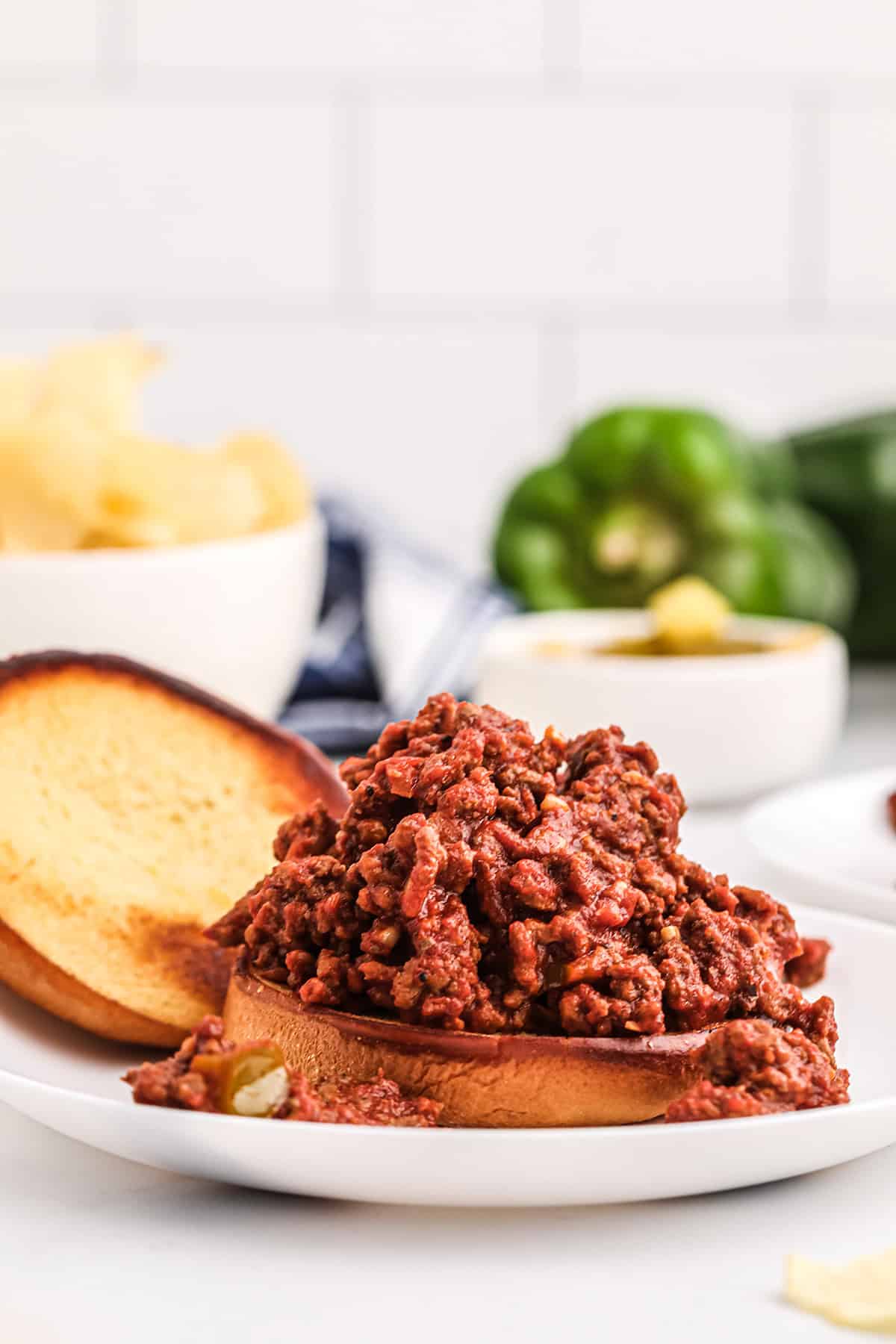 An open-faced sloppy joe sandwich with seasoned ground beef on a toasted bun, served on a white plate with chips and green bell peppers in the background.