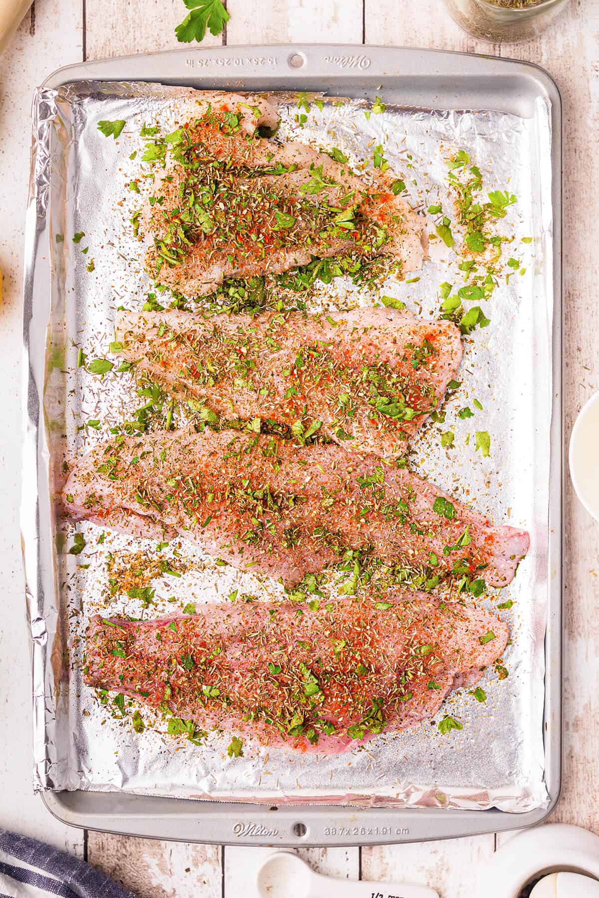 Catfish fillets on a foil-lined pan with herb and seasoning mixture sprinkled over top