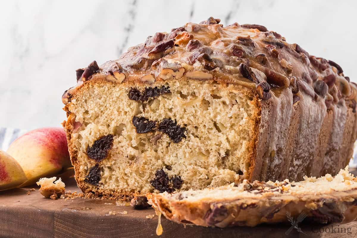 A loaf of fruit and nut bread sits on a wooden surface, with one slice cut and partially removed. The bread contains visible raisins and nuts. There are two apple slices next to the loaf.