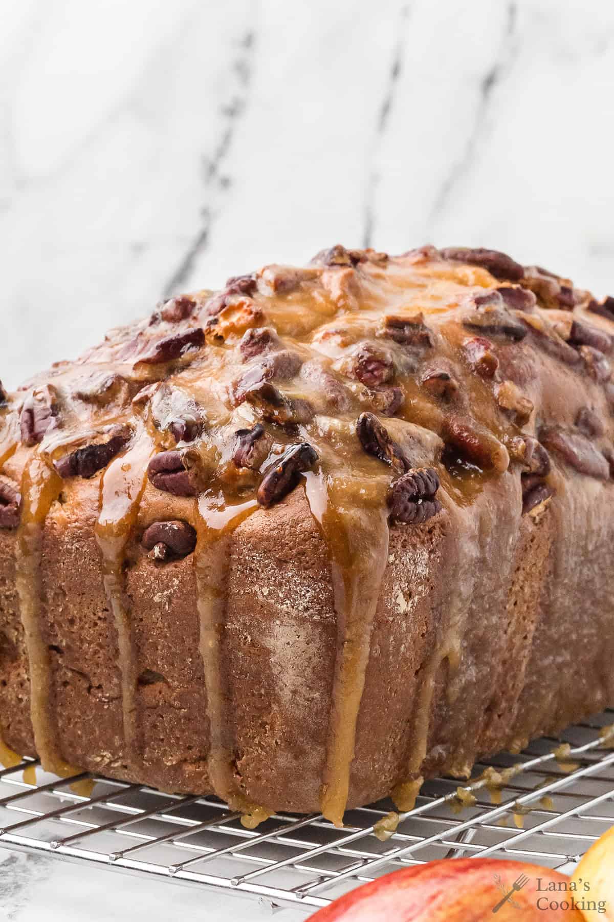 A loaf of bread topped with chopped pecans and caramel glaze sits on a wire cooling rack with a white marble background.