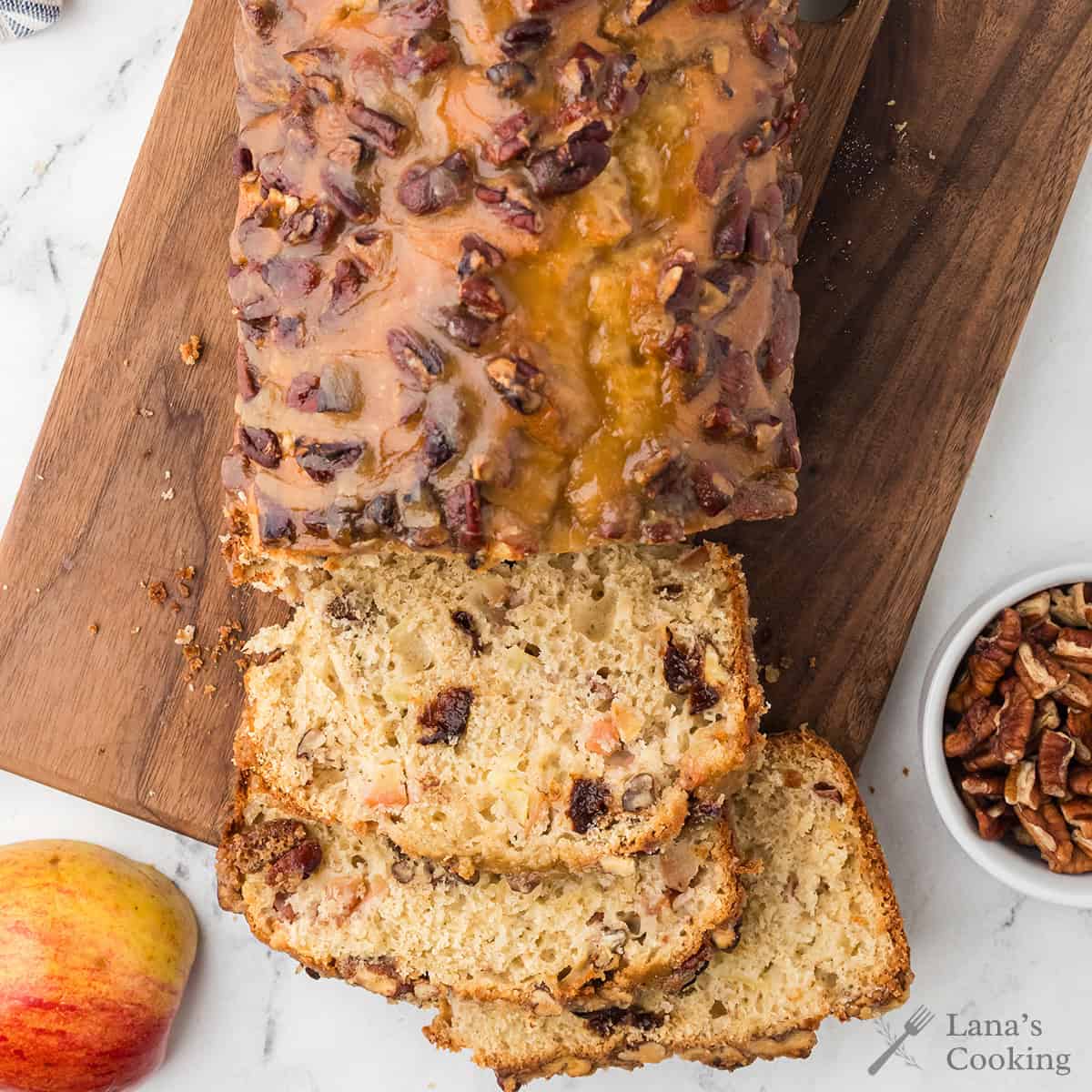 A glazed loaf of bread with pecans on top sits on a wooden cutting board; several slices are cut. An apple and a small bowl of pecans are nearby on a marble surface.