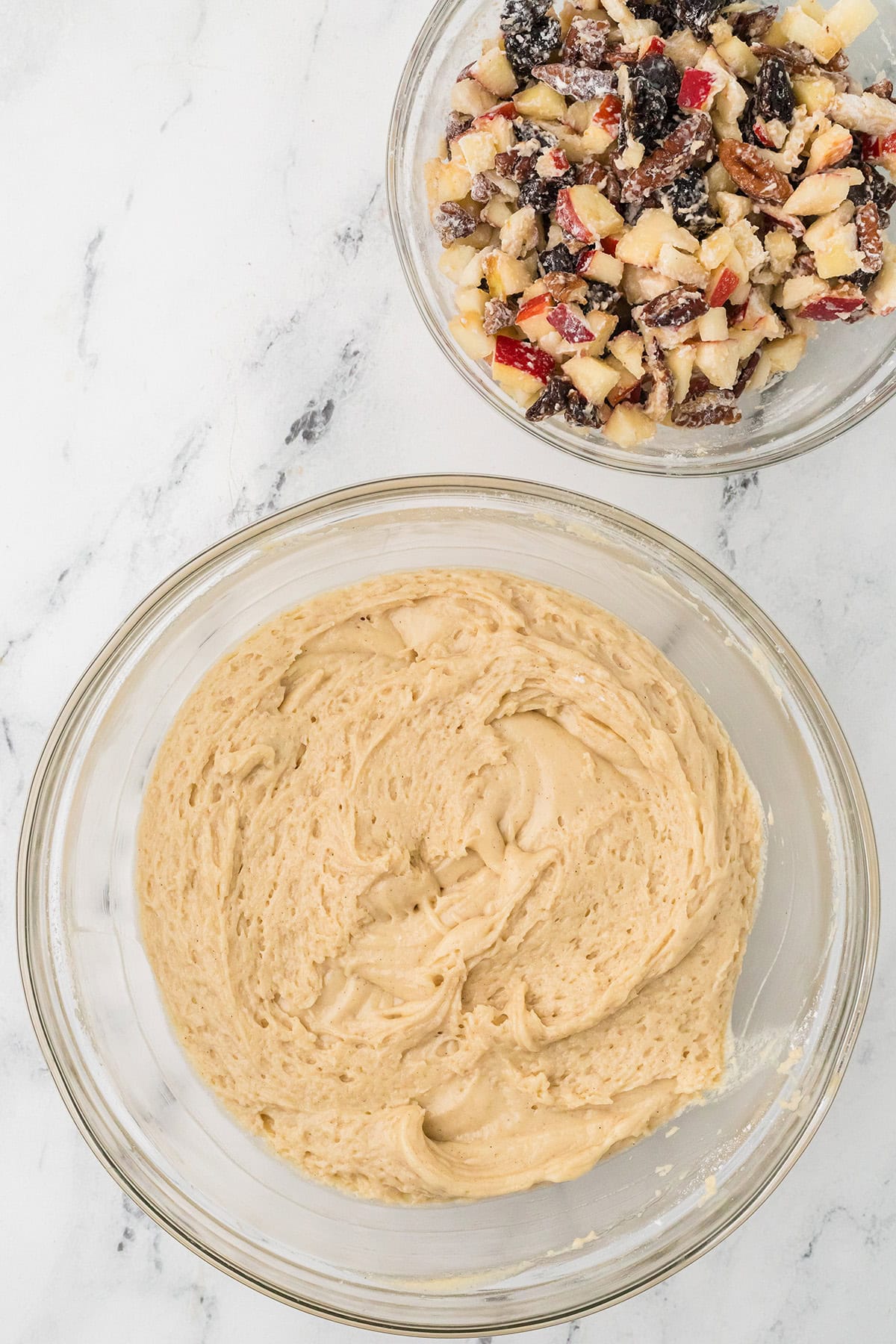 Two glass bowls on a marble surface: one contains a creamy batter, and the other holds a mixture of chopped apples, nuts, and dried fruit.