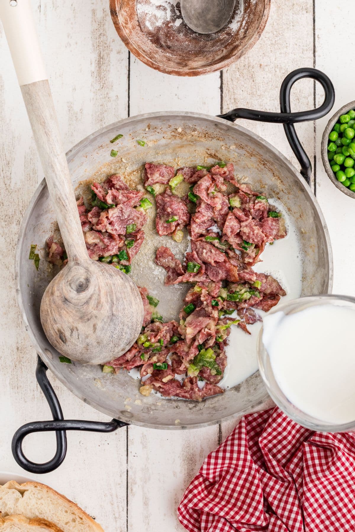 Milk being poured into the beef, flour, onion, and butter mixture and being stirred with a wooden spoon.