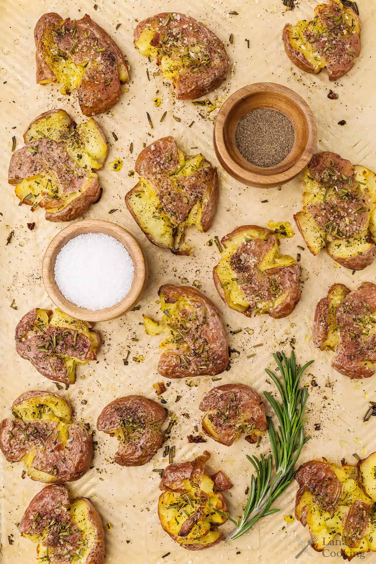 Smashed roasted potatoes with herbs on parchment, with small bowls of salt, pepper, and fresh rosemary.