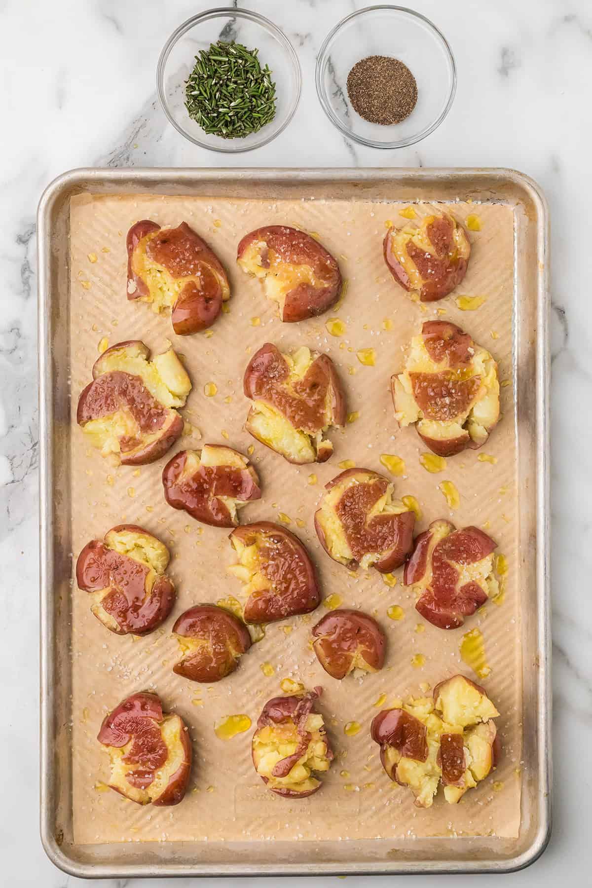 Smashed red potatoes on a baking sheet with bowls of chopped herbs and ground black pepper nearby.