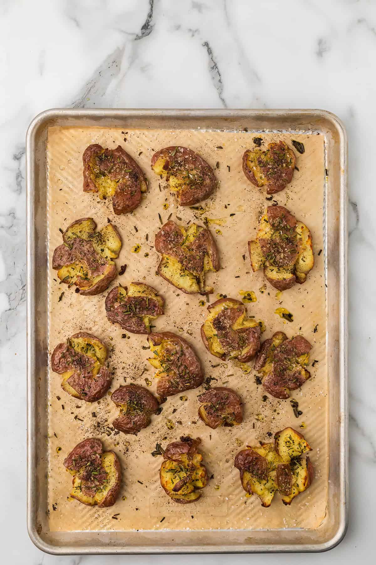 Baked smashed potatoes with herbs on a parchment-lined baking sheet, set on a marble countertop.