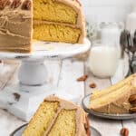 Two slices of yellow cake with caramel frosting and pecans on plates, with the rest of the cake in the background.