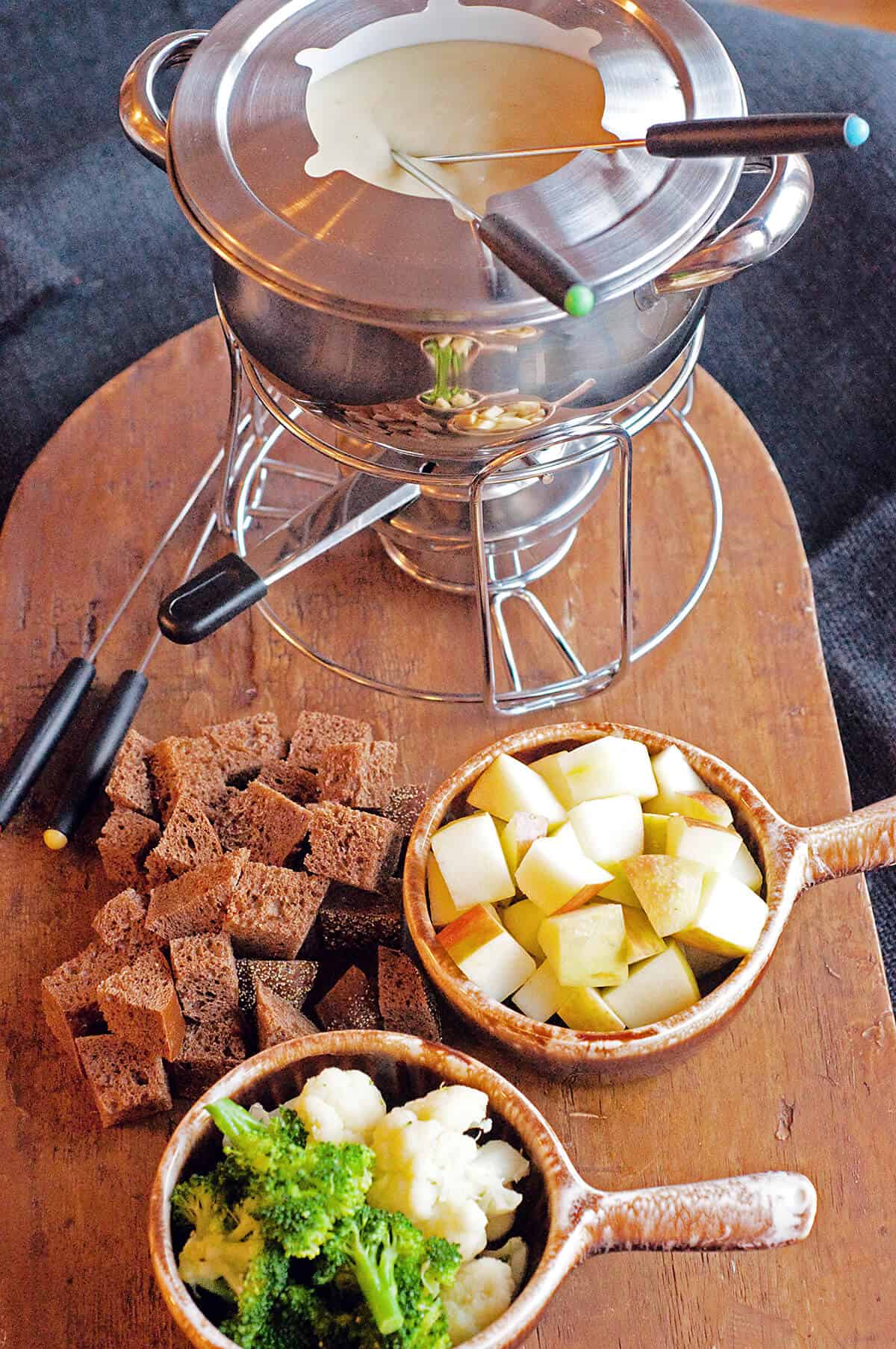 Fondue pot filled with cheese fondue and bread, fruit, and veggie dippers alongside.