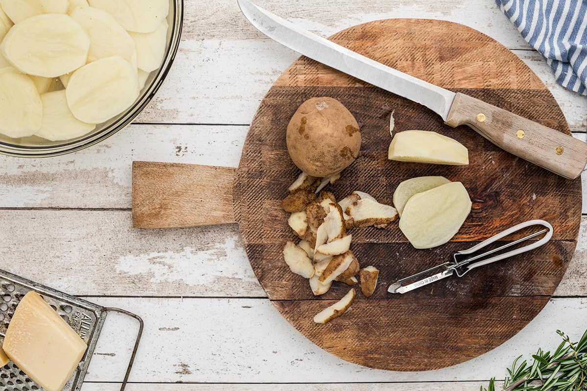 Potatoes, a knife, and a vegetable peeler on a cutting board.