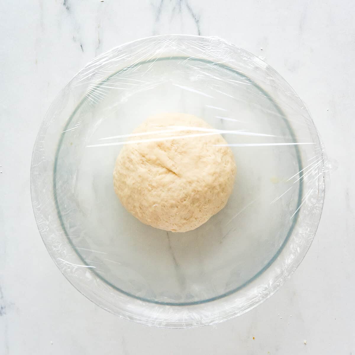 Dough ball in a mixing bowl covered with plastic wrap.