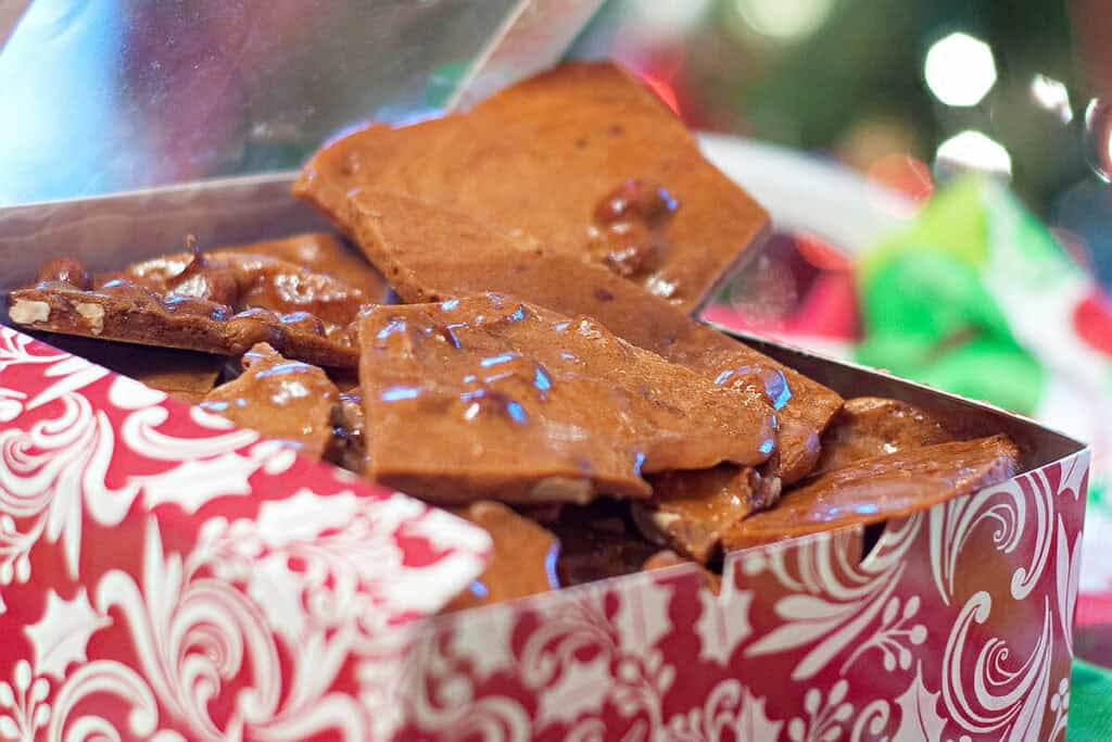 A festive box filled with pieces of nut brittle, with a blurred holiday background.
