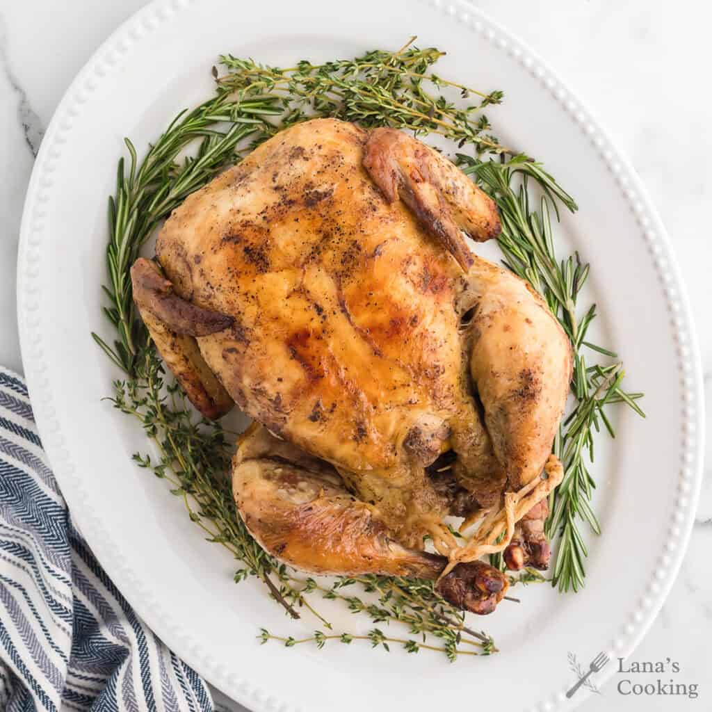 Whole roasted chicken surrounded by fresh herbs on a serving plate.