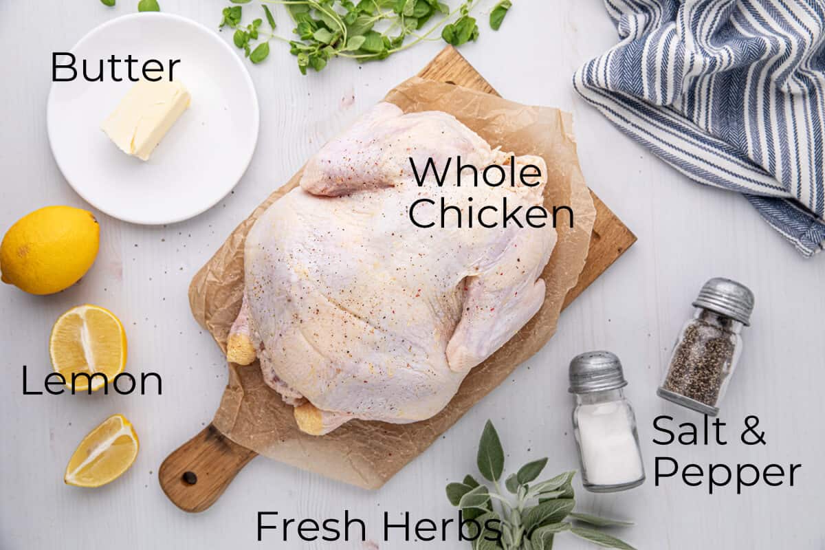 A raw whole chicken on a wooden board surrounded by butter on a plate, lemon halves, fresh herbs, a salt shaker, a pepper shaker, and a striped towel. Each ingredient is labeled with text.