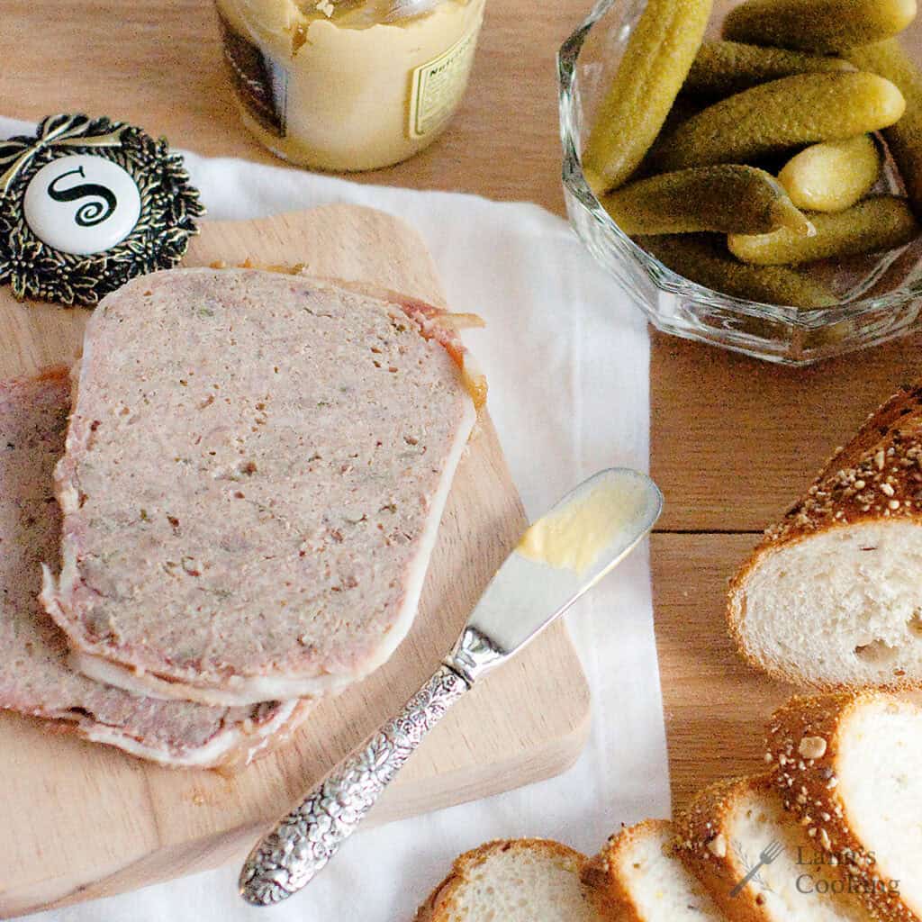 Slices of terrine on a cutting board with bread, gherkins, mustard, and a butter knife nearby.