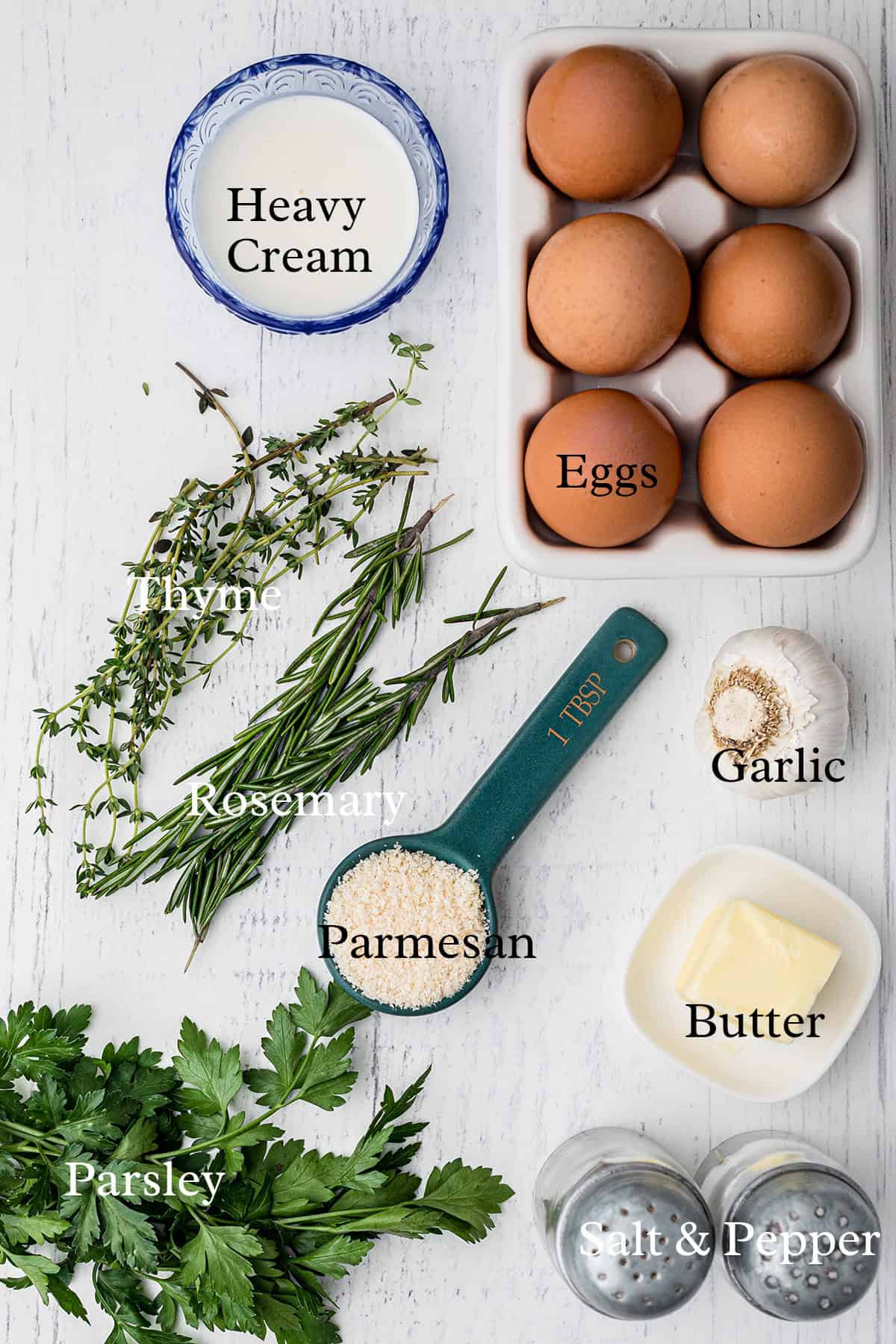 Top-down view of eggs, heavy cream, garlic, butter, herbs, Parmesan cheese, and salt & pepper on a white surface.