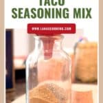 A glass jar filled with layered taco seasoning mix sits on a counter; text reads Homemade Taco Seasoning Mix.
