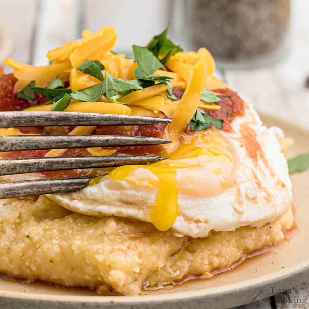 A fork cuts into a fried egg on cheesy grits, topped with salsa, shredded cheese, and parsley.