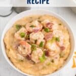 A bowl of shrimp and grits topped with green onions, set on a table with gold utensils and a napkin.